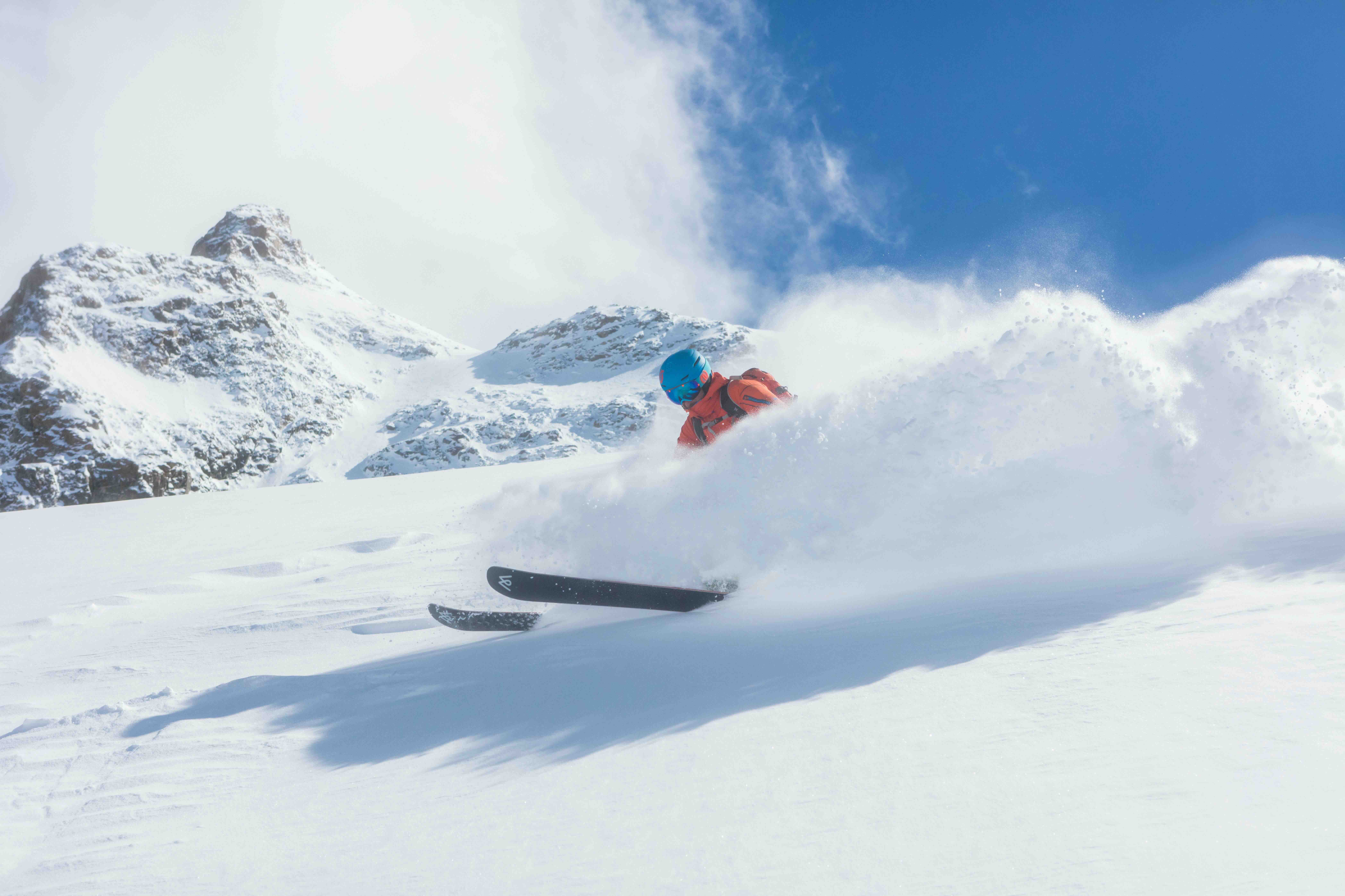A skier in an orange jacket carving through fresh powder snow below rocky, snow-covered mountain peaks