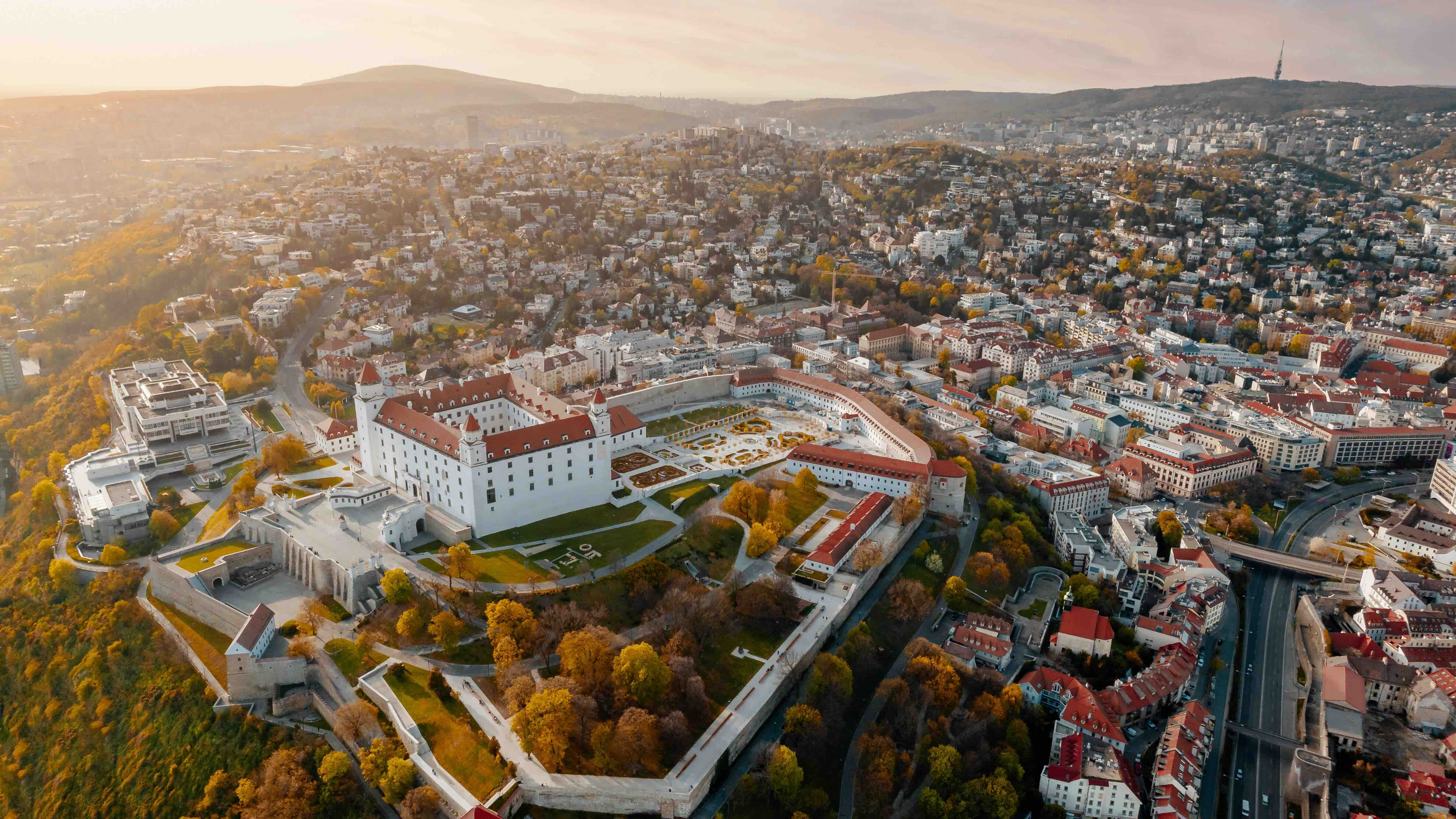Aerial view of Bratislava at golden hour, with the white four-towered Bratislava Castle and its baroque gardens on the hill above the city, the rooftops and wooded hills spreading beyond