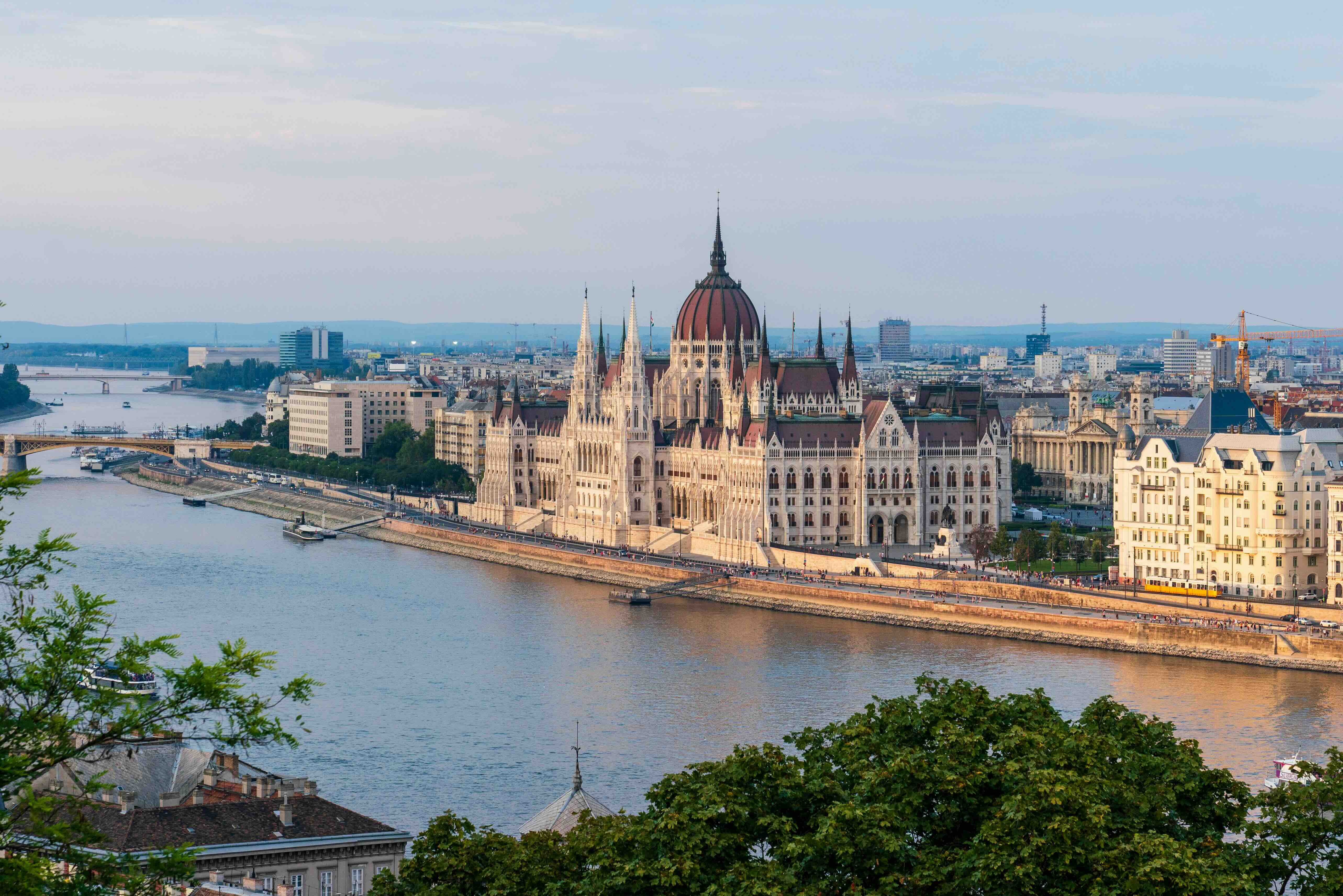 The neo-Gothic Hungarian Parliament building on the Danube embankment in Budapest, its red dome and spires above the Pest riverfront, with a yellow tram passing on the promenade at dusk