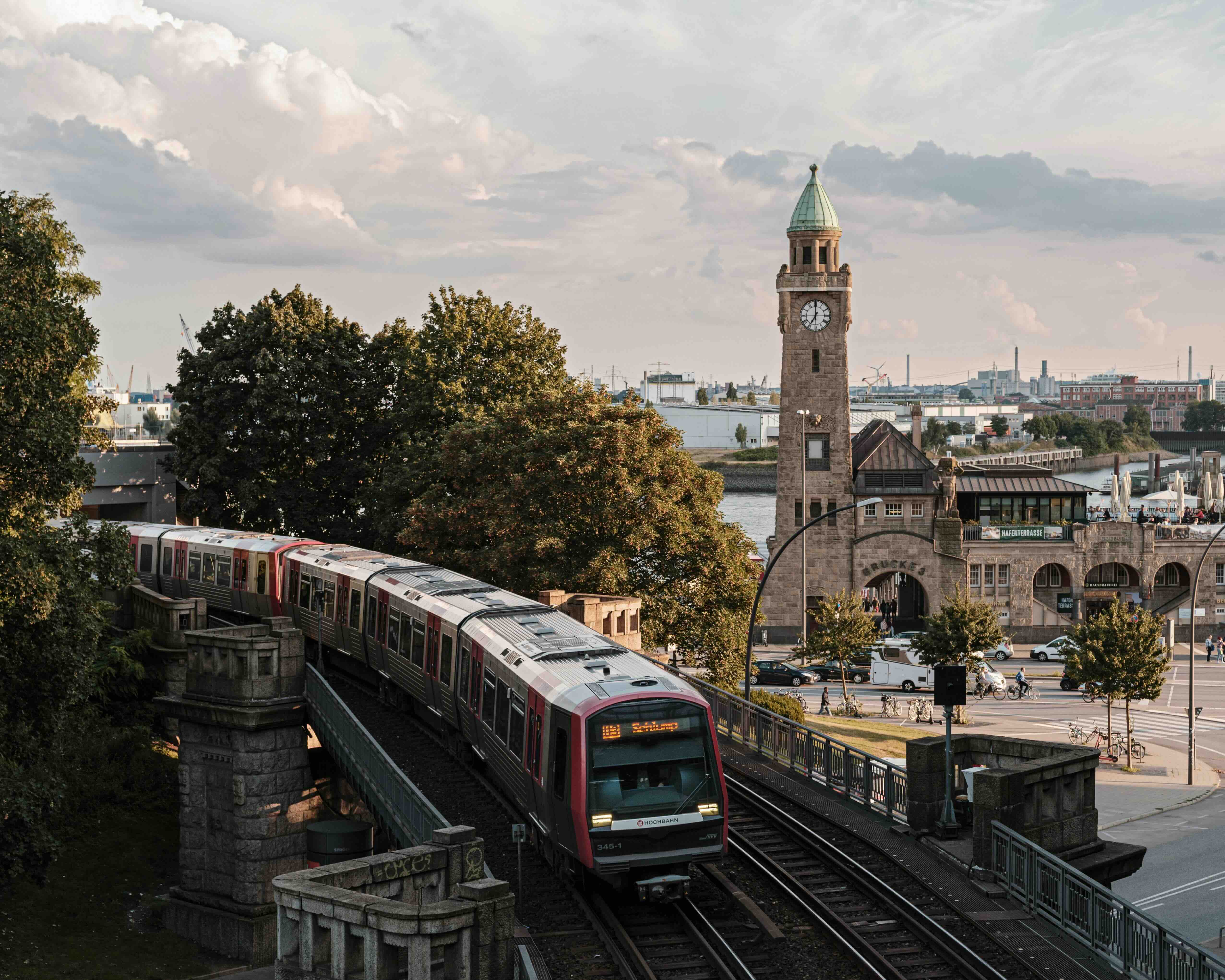 A red and white Hamburg U-Bahn train crossing the elevated viaduct by the harbour at Landungsbrücken, with the historic clock tower of the St Pauli Landungsbrücken and the port on the Elbe beyond