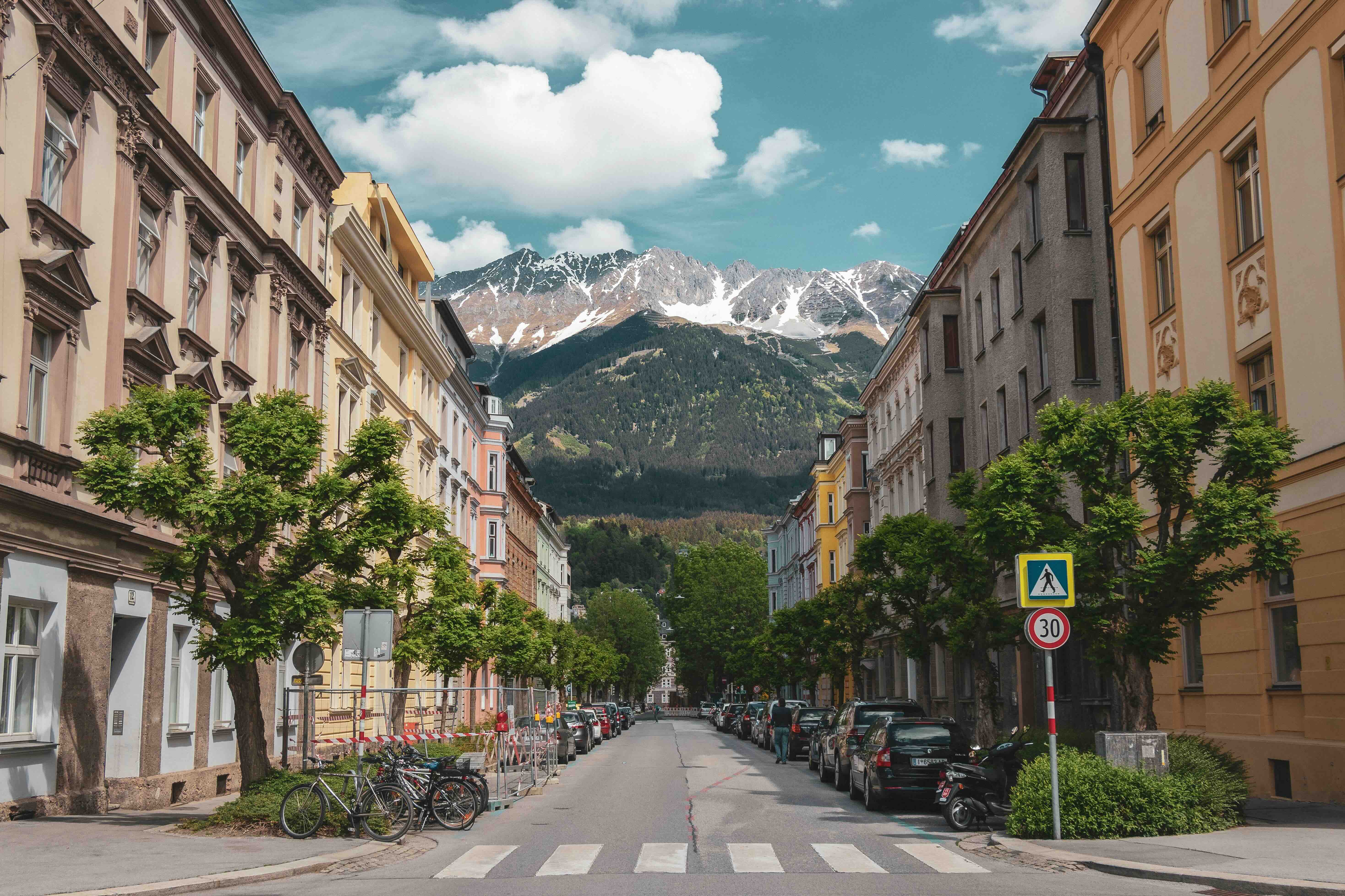 A tree-lined street of pastel townhouses in Innsbruck framing the snow-capped peaks of the Nordkette mountain range rising steeply beyond the city under a blue alpine sky