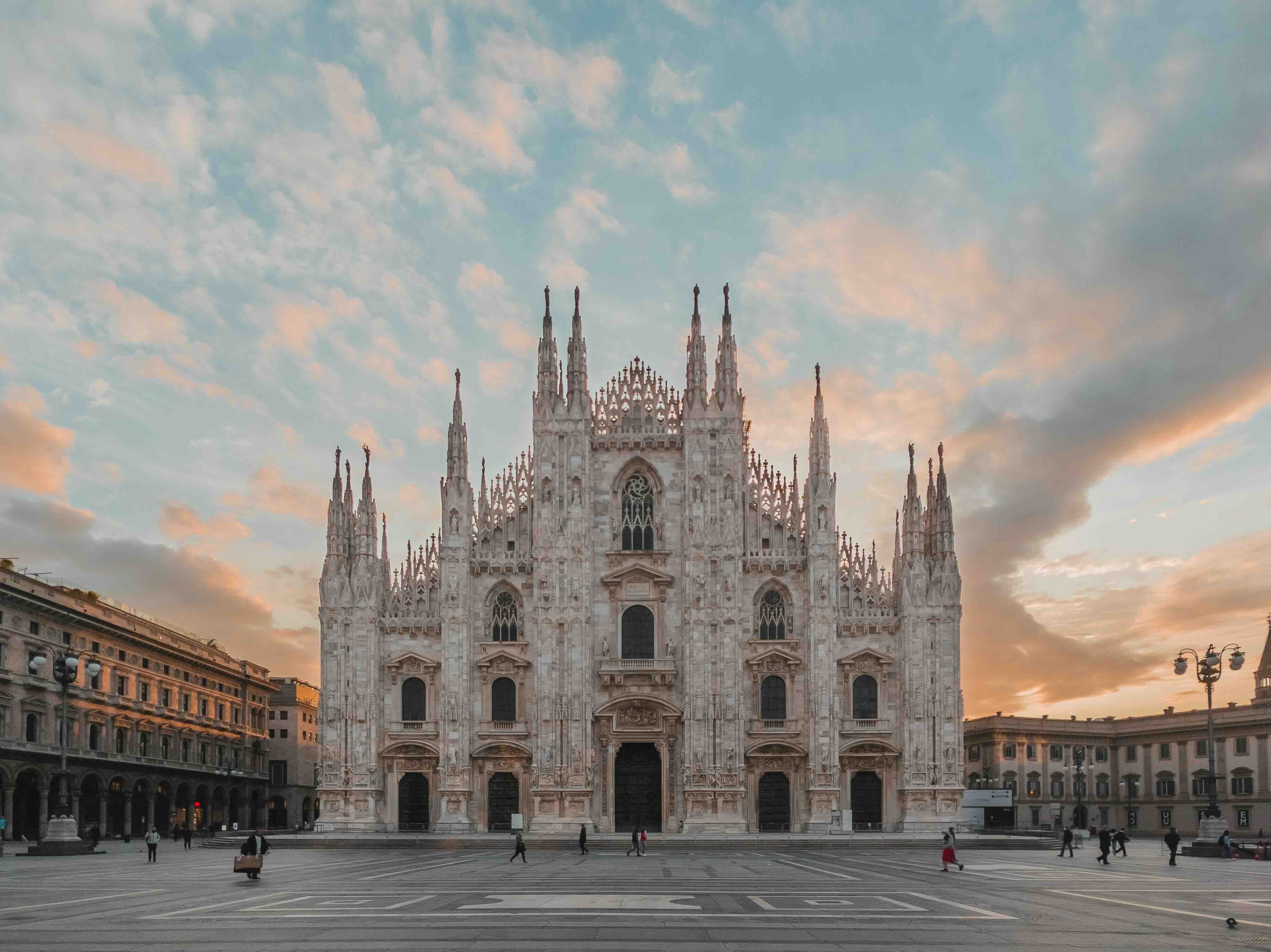 The white marble Gothic facade and spires of Milan's Duomo cathedral rising over the empty Piazza del Duomo at dawn under a soft pink and orange sky