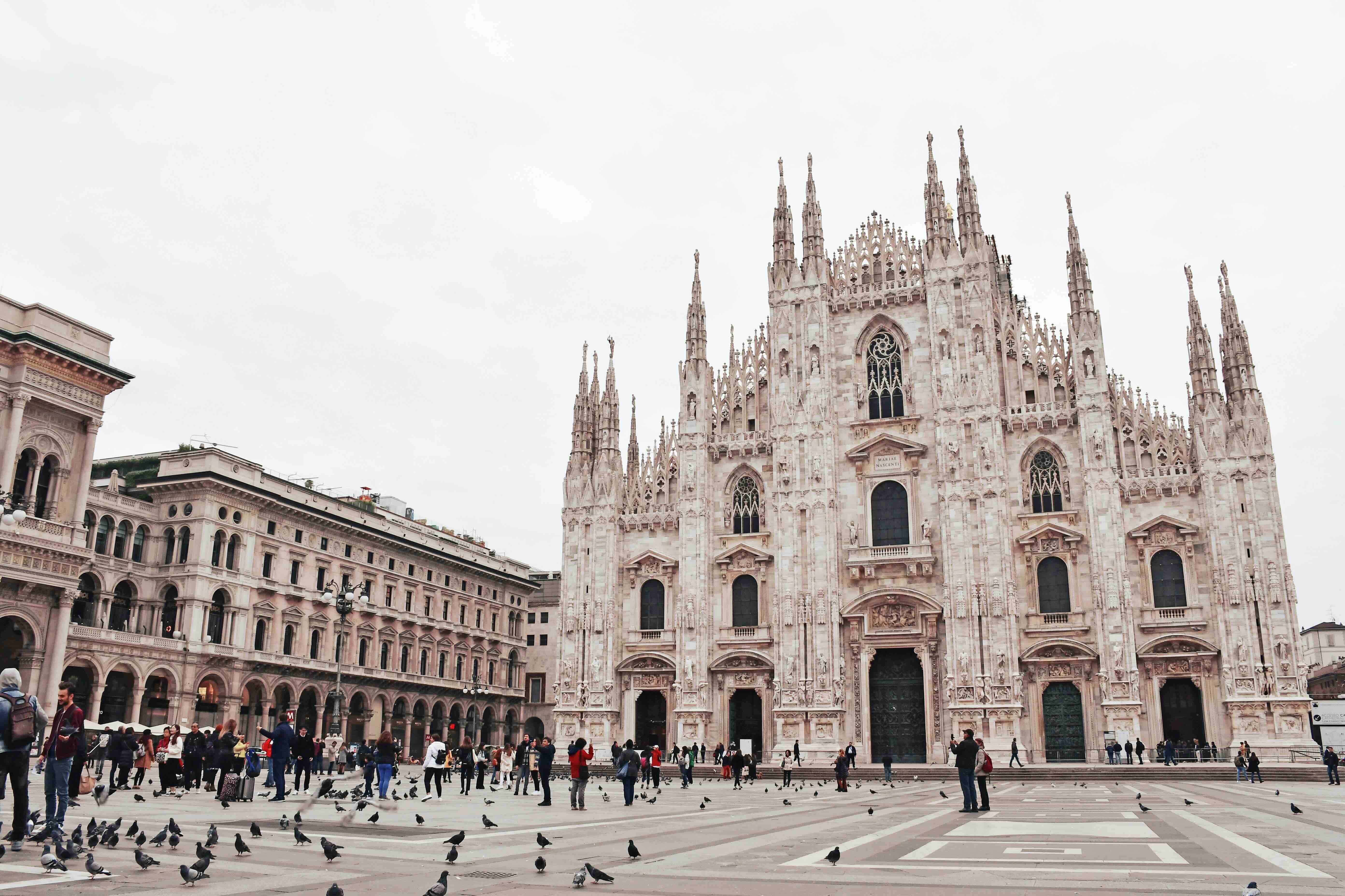 The white marble Gothic facade and spires of the Duomo di Milano rising over the Piazza del Duomo, with people and pigeons crossing the square