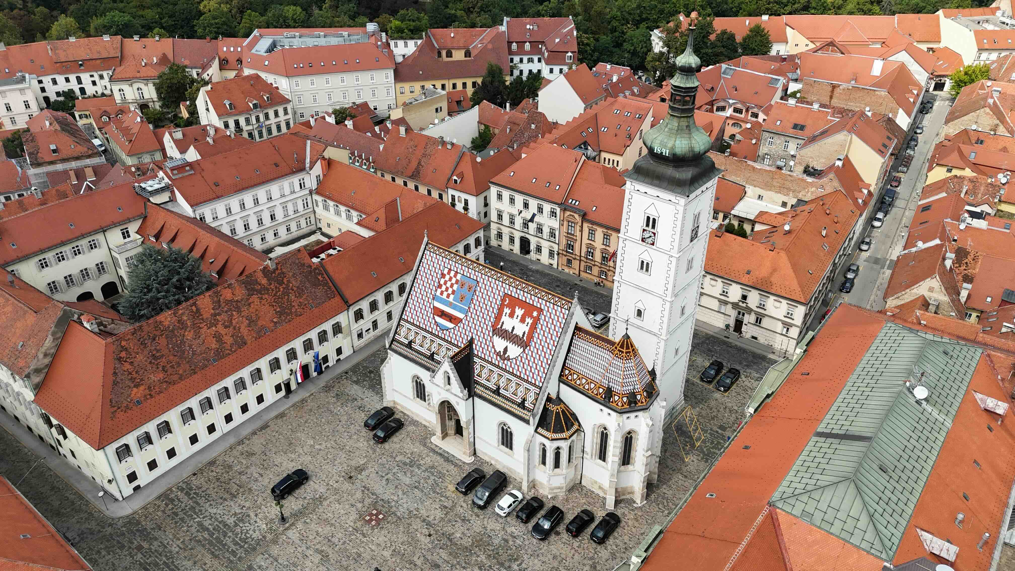 An aerial view of St Mark's Church in Zagreb's Upper Town, its steep tiled roof bearing the coats of arms of Croatia and the city of Zagreb, beside its white bell tower with a green dome, ringed by the red-roofed buildings of the old town